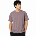A man wearing a plain crew-neck t-shirt in a muted dusty mauve or taupe color, paired with black pants, photographed against a solid white background.