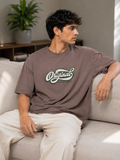A man sitting on a light beige sofa wearing a brownish-gray oversized t-shirt with a white and green 'Original' graphic on the chest, paired with off-white pants.
