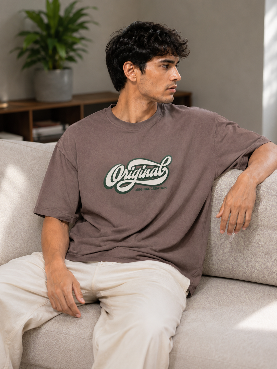 A man sitting on a light beige sofa wearing a brownish-gray oversized t-shirt with a white and green 'Original' graphic on the chest, paired with off-white pants.