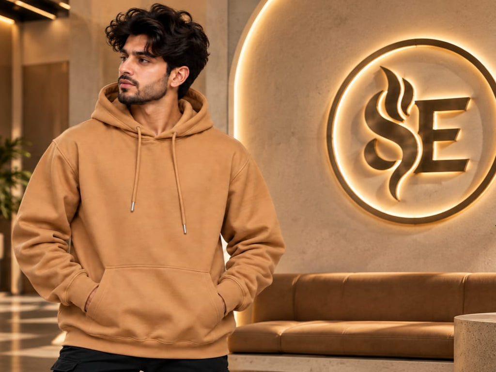 A man wearing a camel brown hoodie stands in a modern interior lobby with a large illuminated 'SE' logo on the wall behind him.