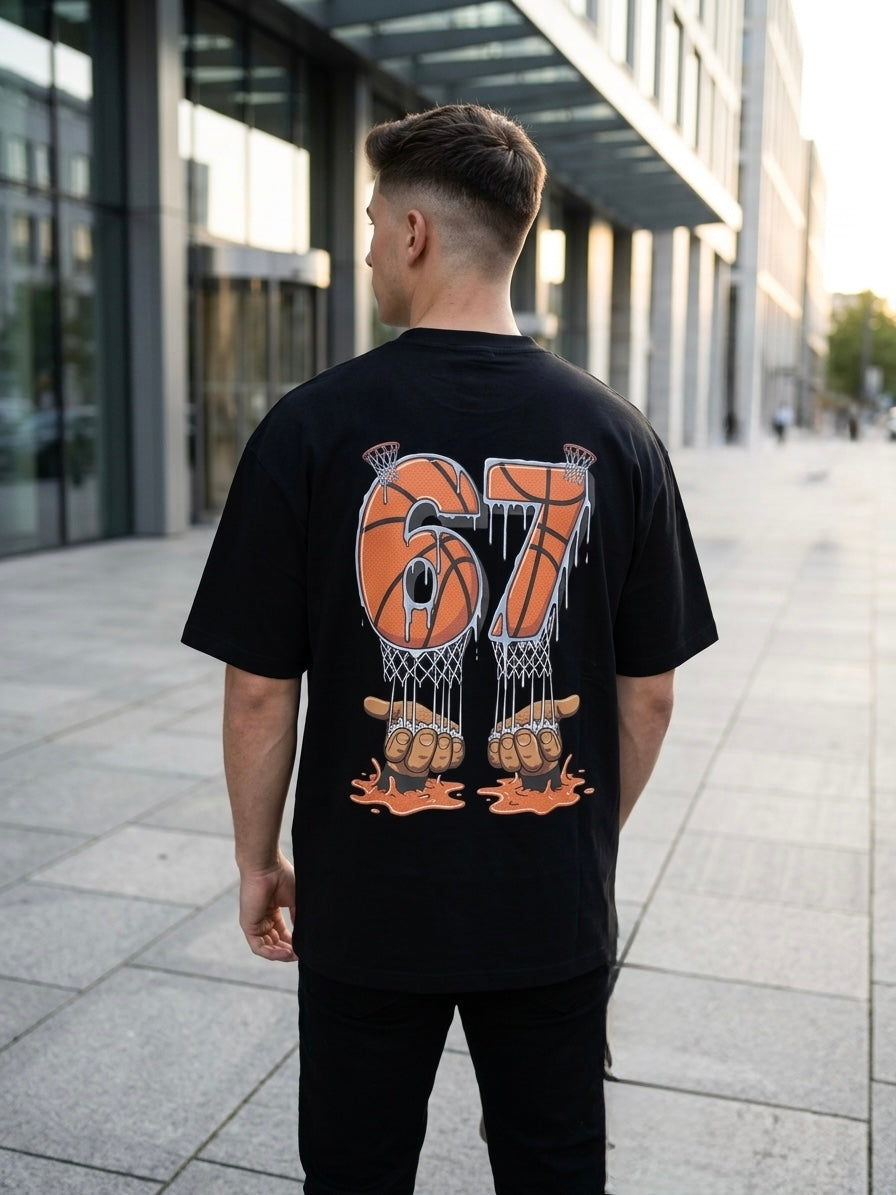A man viewed from behind wearing a black t-shirt with a large orange and white graphic on the back featuring the number 67 and basketball-themed design, standing on a paved walkway in front of modern glass buildings.