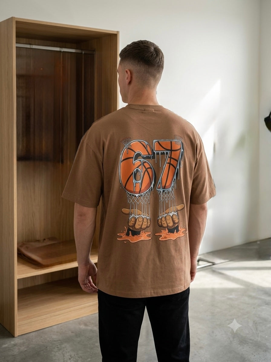 A man viewed from behind wearing a brown oversized t-shirt with a large basketball-themed graphic on the back, standing in a minimalist room with a wooden shelf and concrete floor.